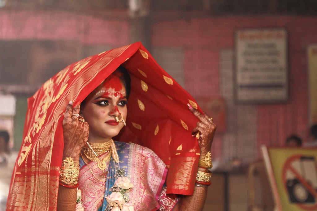 A stunning portrait of a Bengali bride dressed in a red Banarasi saree with traditional sandalwood (Chandan) art on her forehead.